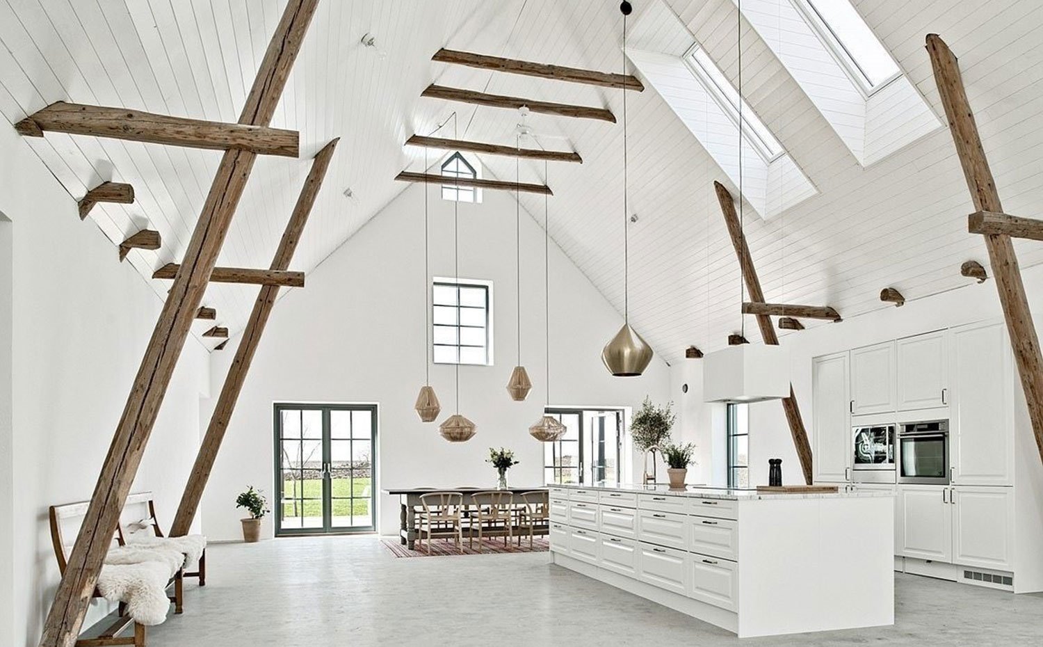 Cocina comedor de gran amplitud con cubierta de madera pintada de blanco, vigas dispuestar horizontal y verticalmente, mobiliario en blanco y grandes luminarias suspendidas 