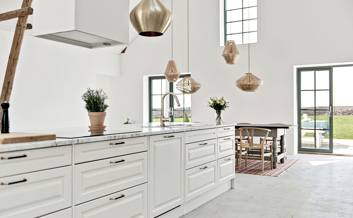 Cocina con mobiliario en blanco, tiradores cromados, encimera de mármol blanco, luminaris suspendidas a gran altura y mesa y sillas de comedor en madera