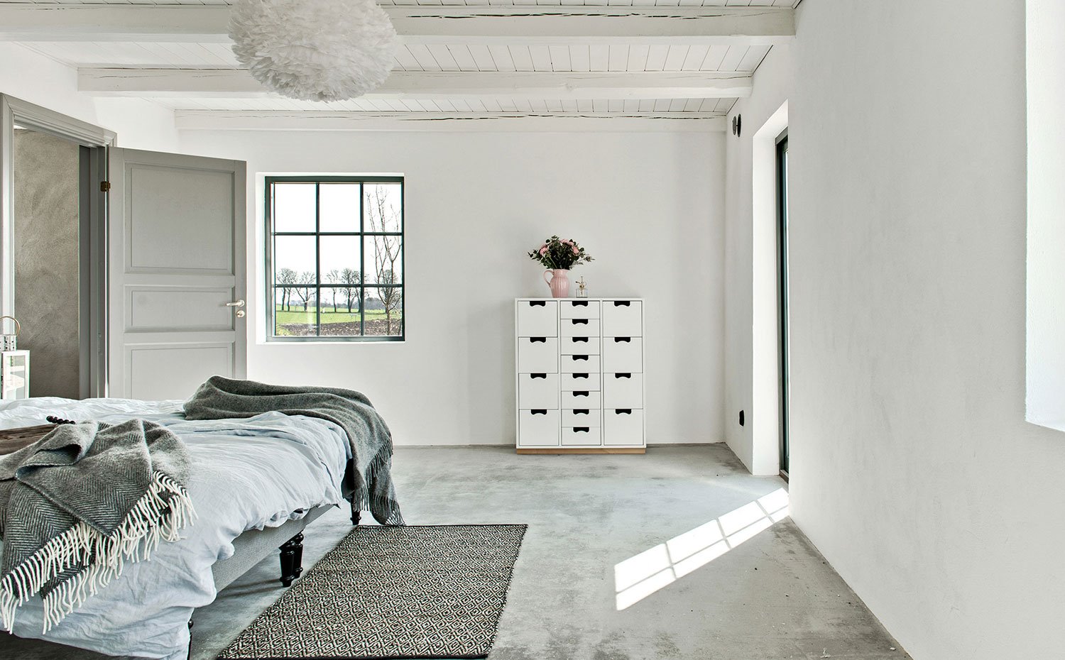 Dormitorio con cómoda en blanco y tiradores negros, puerta y marco en gris, vigas y techo de madera enblanco, suelo de hormigón