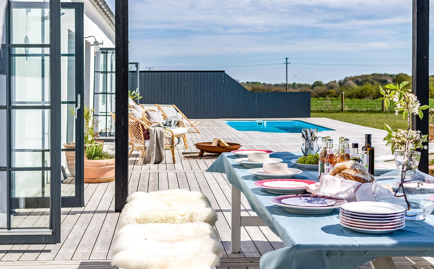 Mesa de comedor exterior con asientos revestidos de piel sintética blanca, entarimado de madera y piscina al fondo
