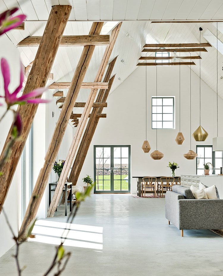 Vigas de madera de suelo a techo junto a paramentos blancos, techo a dos aguas de madera pintada de blanco, sofá en gris