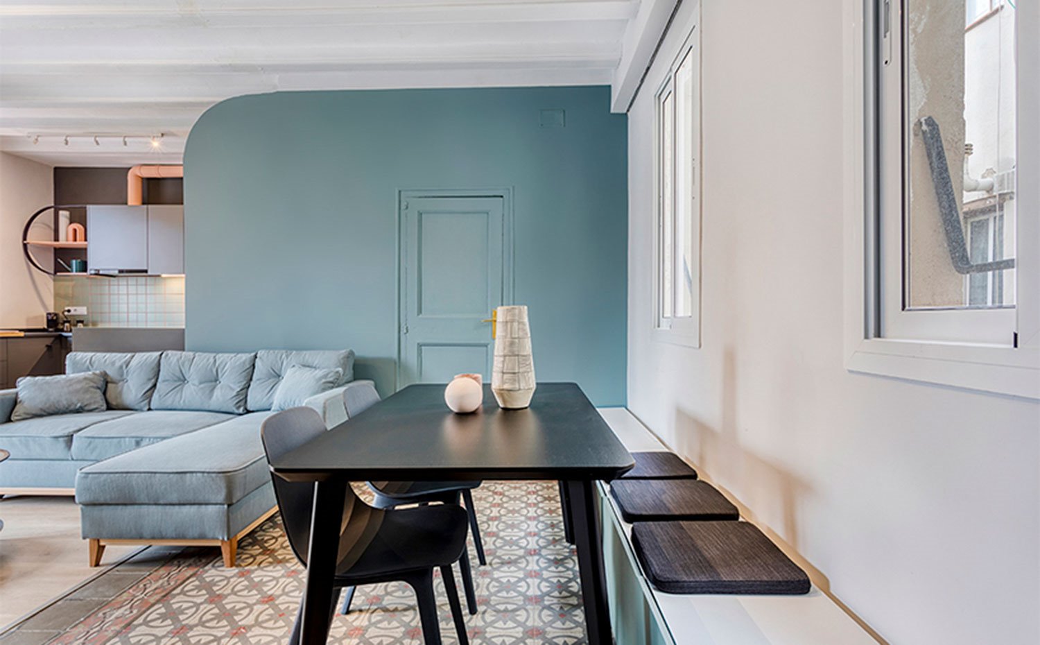 Comedor con mesa y sillas de madera oscura y módulos bajos que actúan como banco con cojines en tonos grises