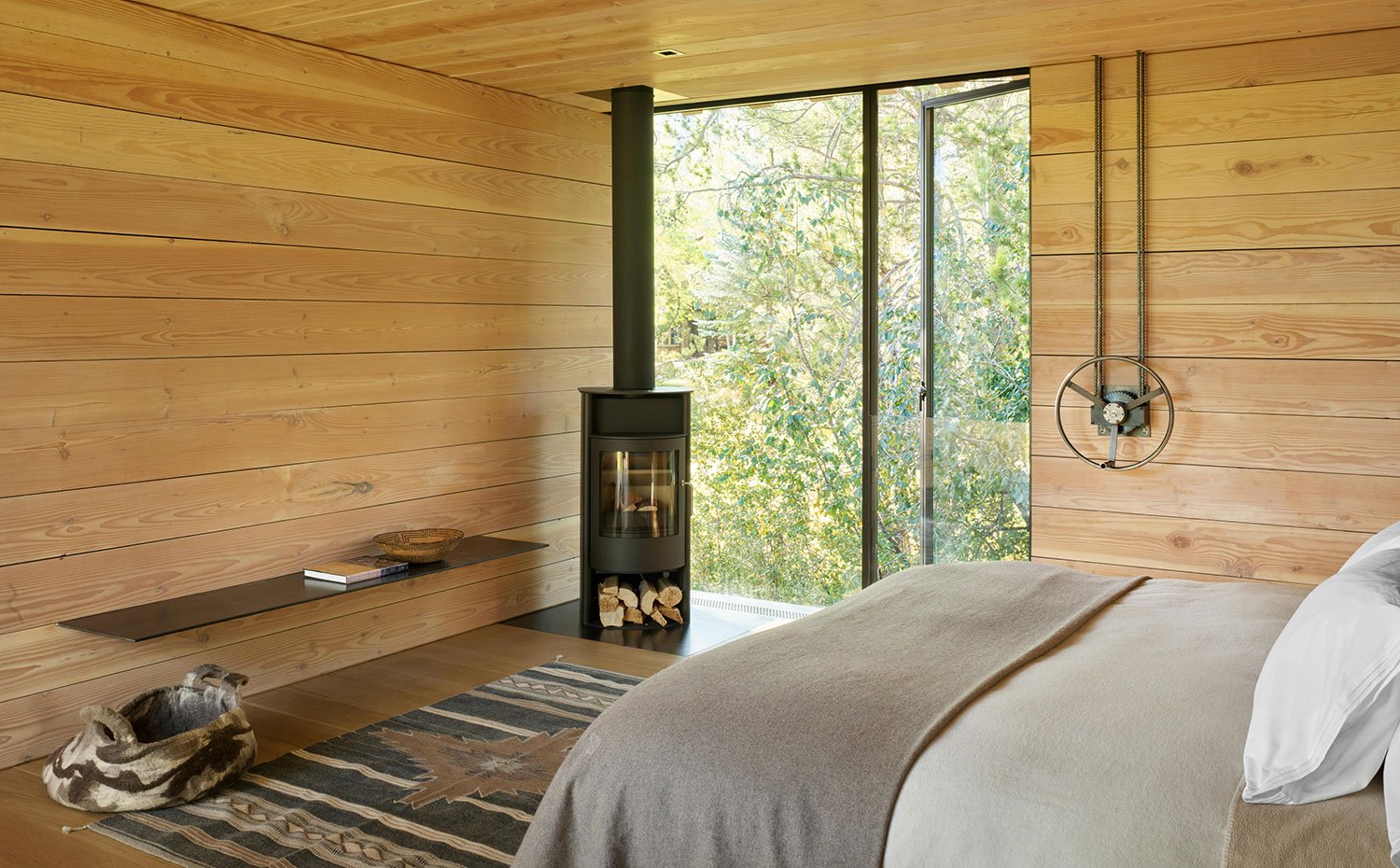 Dormitorio con chimenea en esquina junto a apertura a zona boscosa, paredes de madera con banco suspendido en acero