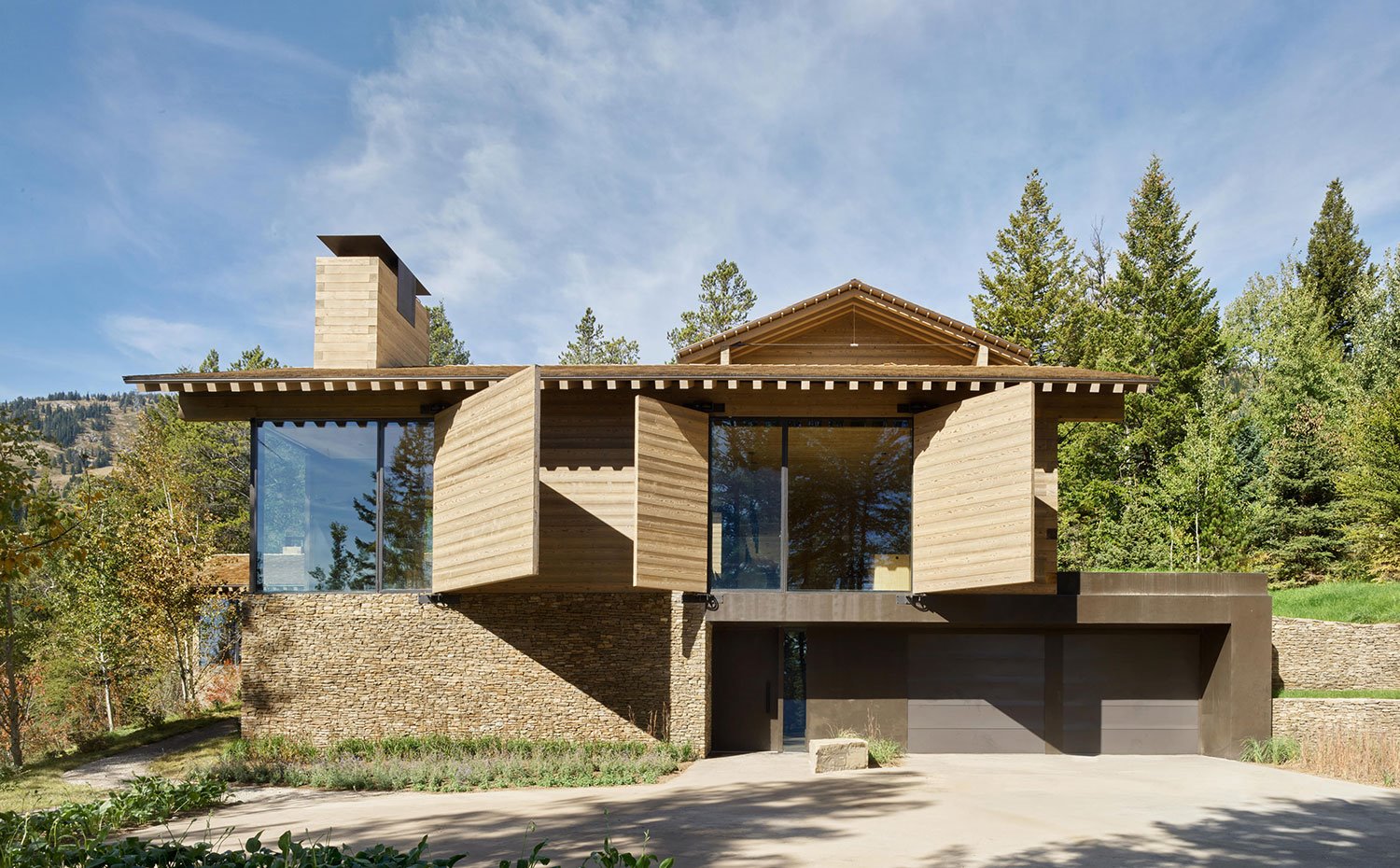 Fachada frontal de vivienda con estructura de piedra, cristal y madera, entre pinos y zona boscosa