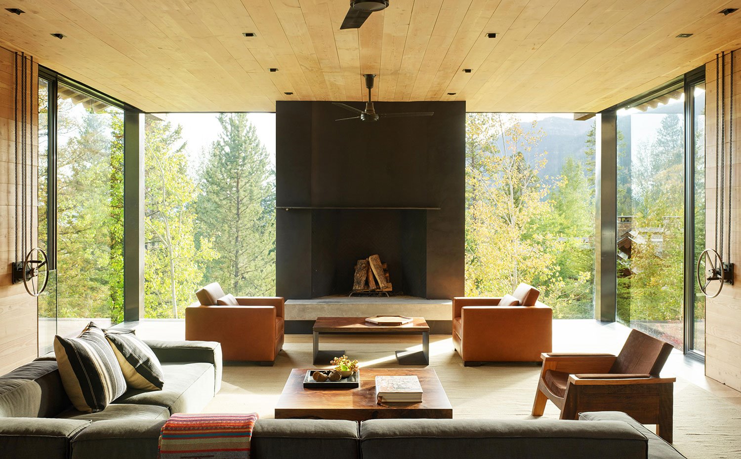 Salón con chimenea central, cerramientos acristalados, sillones en marrón, sofás en gris, butaca y mesa en madera