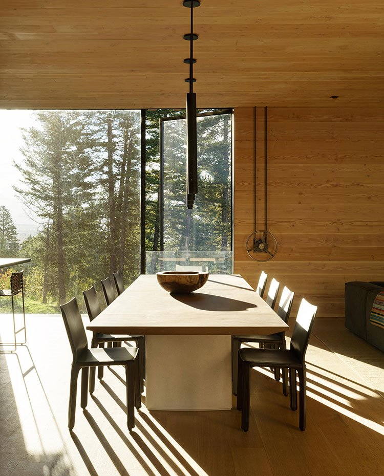 Zona de comedor con mesa de madera y sillas de piel marrón, luminarias suspendidas y amplia apertura acristalada hacia exterior
