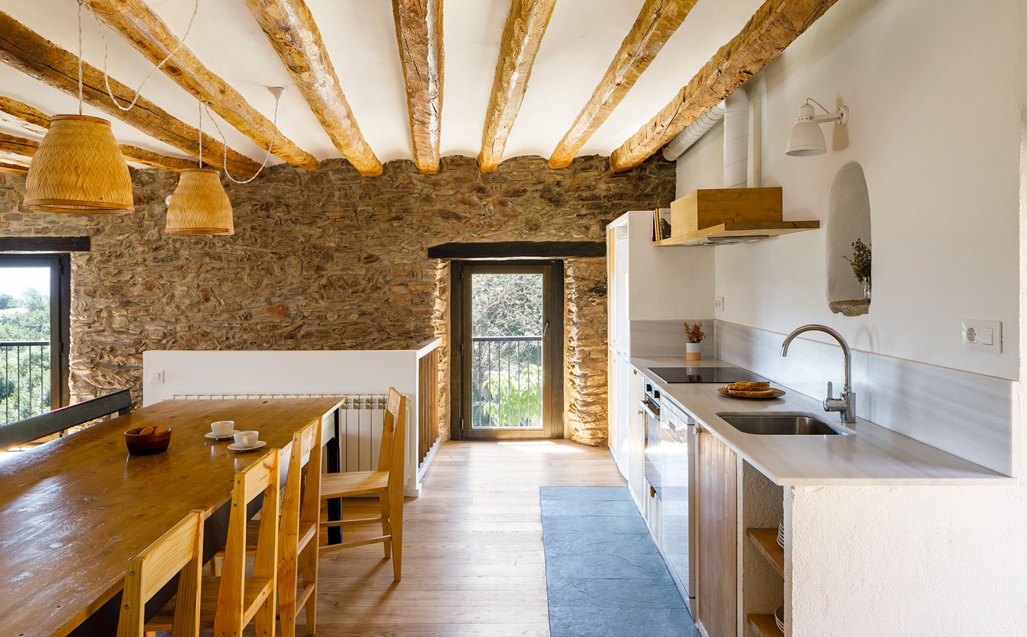 Comedor y cocina abierta en un mismo espacio con vigas de madera, luminarias suspendidas de fibras naturales y pared de piedra