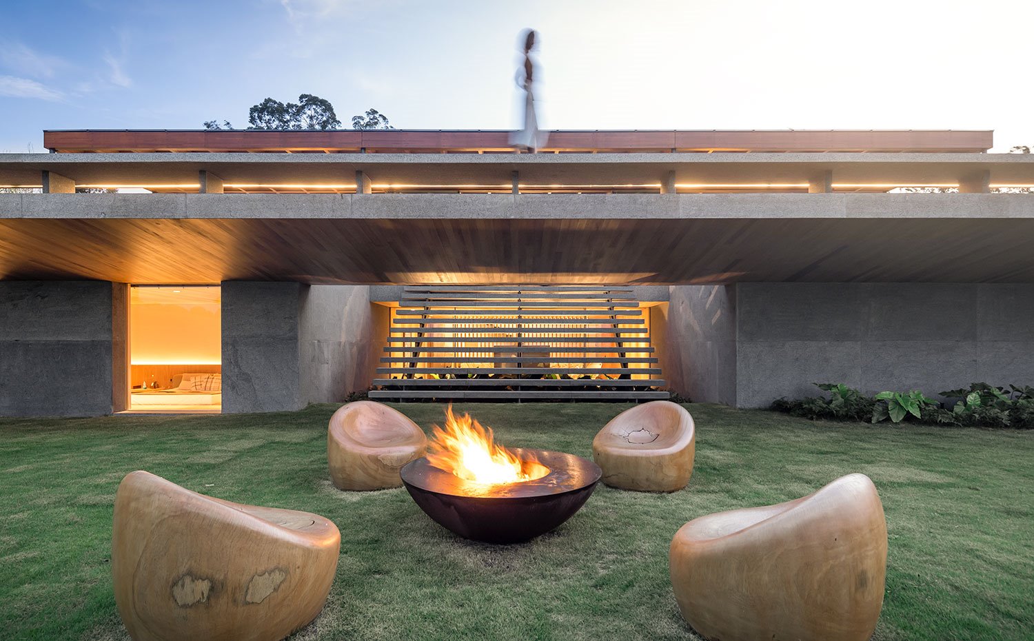 Chimenea encendida sobre jardín, butacas de madera en círculo frente a vivienda al atardecer