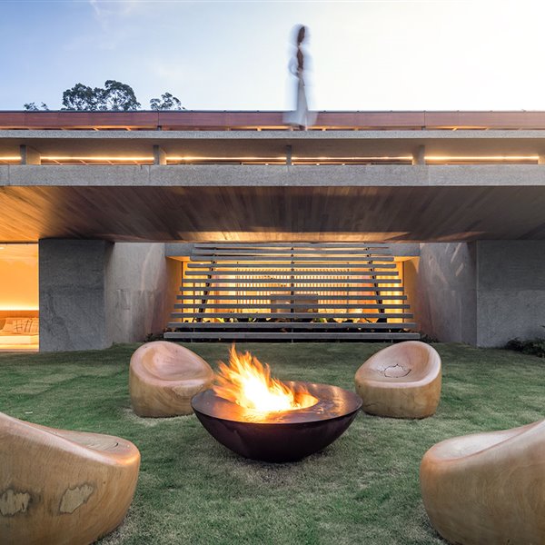 Chimenea encendida sobre jardín, butacas de madera en círculo frente a vivienda al atardecer