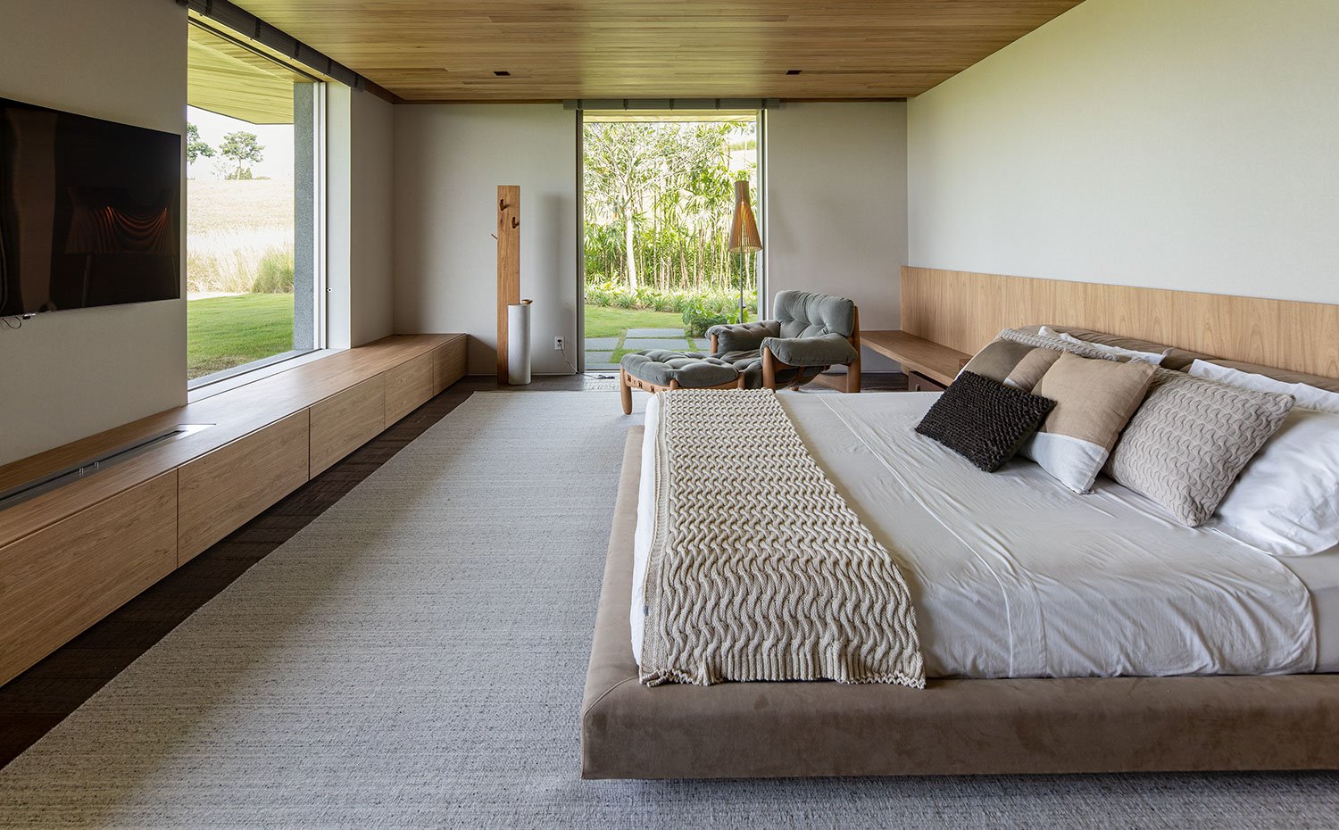 Dormitorio con estructura de cama tapizada en marrón, cabecero y mueble frontal hecho a medida en madera natural, butaca y reposapies de madera y tapizado en verde