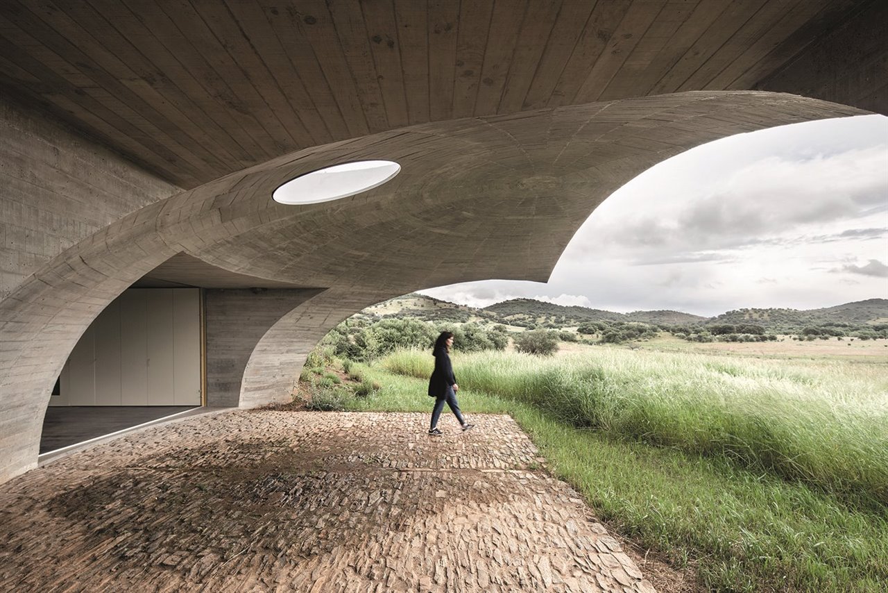 Un espectacular medio arco de hormigón perforado por una claraboya circular se proyecta desde el terreno para generar el amplio porche pavimentado con piedra.