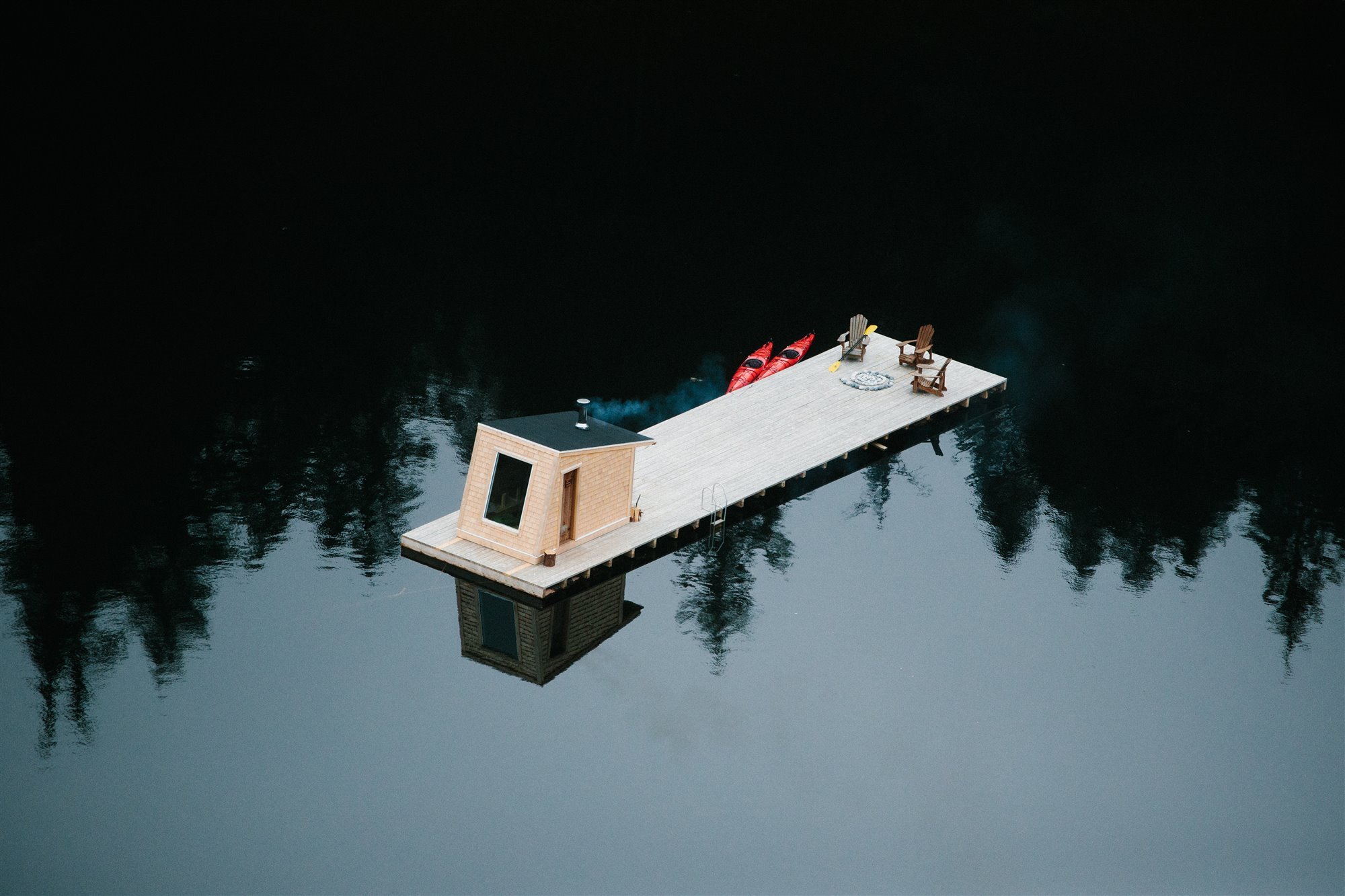 Cabaña moderna con Sauna de madera flotando en mitad de un lago con canoas y tumbonas