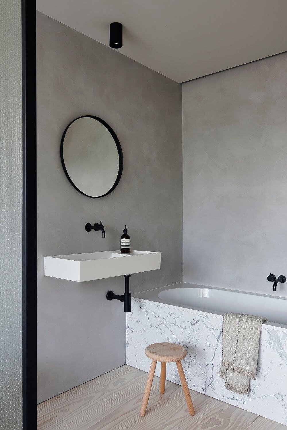 Lavabo blanco suspendido en cuarto de baño gris con grifería negra y bañera de mármol.