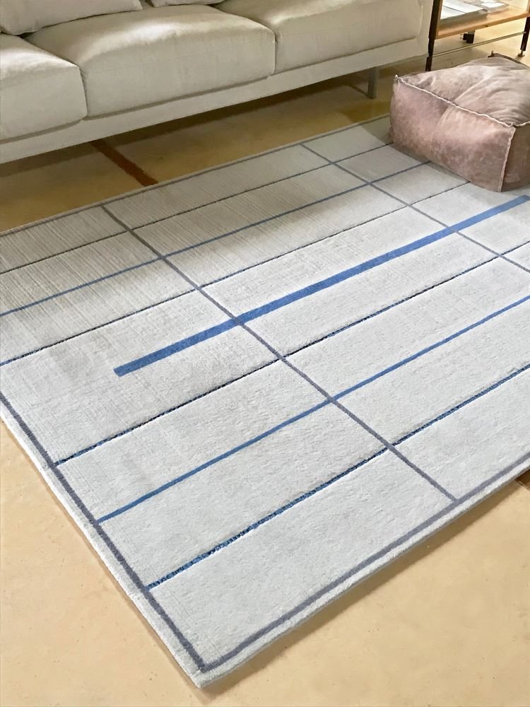  El modelo Raak tiene un fondo gris claro sobre el que las finas líneas grises y azules establecen un diálogo con la importante franja azul que se interrumpe sin llegar al borde, creando una sutil tensión en el conjunto