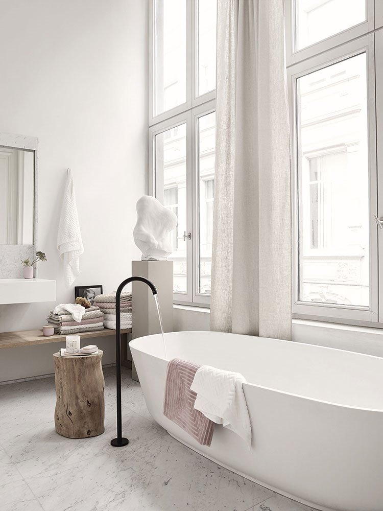 Paredes blancas, detalles de madera, suelo mármol y, surgiendo de él, de una forma casi poética, un grifo negro para la bañera exenta.
