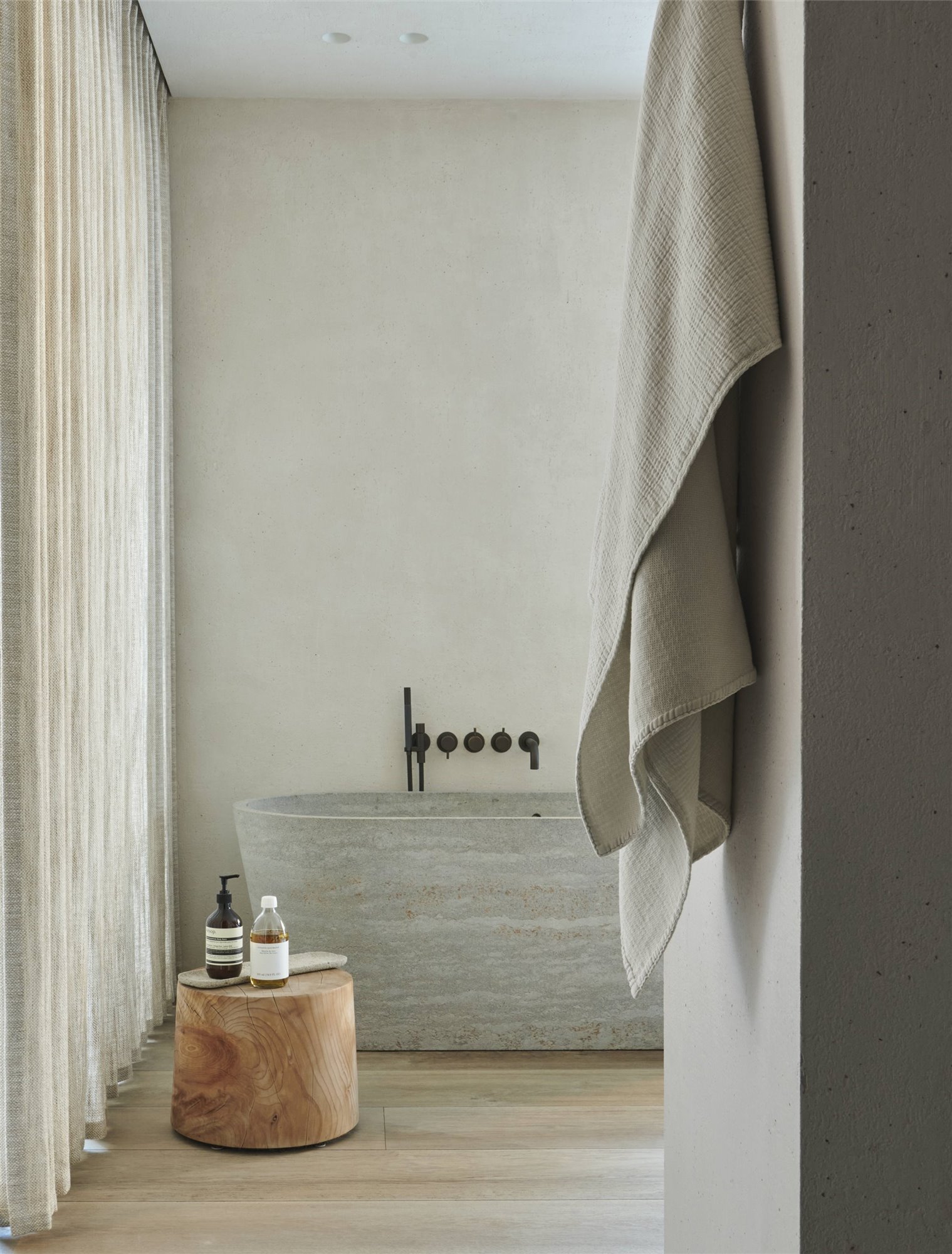 Baño con bañera de piedra con grifos negros y taburete de madera.