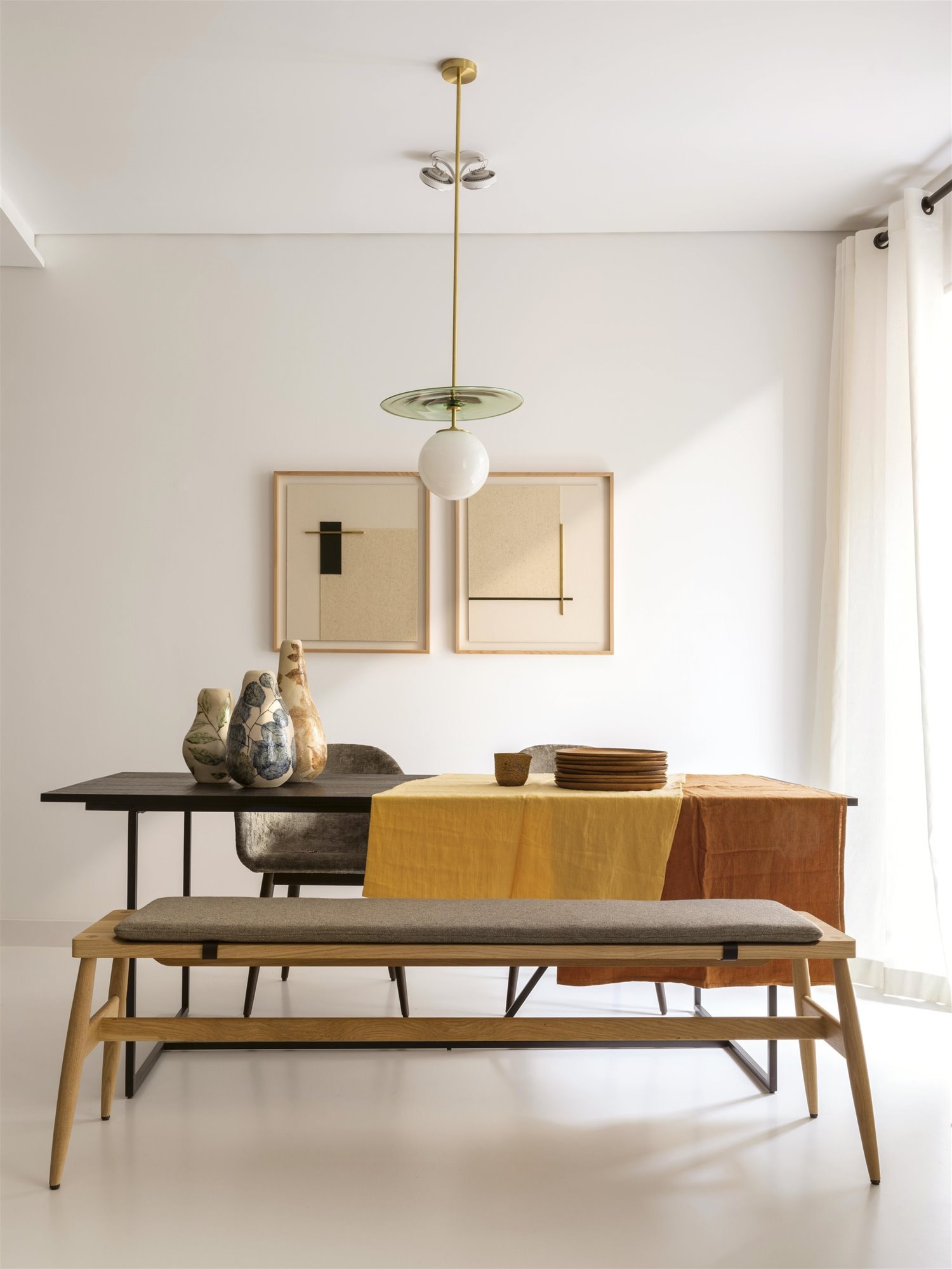 mesa comedor rectangular con banco y mantel naranja y amarillo, jarrones de cerámica, platos, lámpara de techo verde y blanca y cuadros abstractos