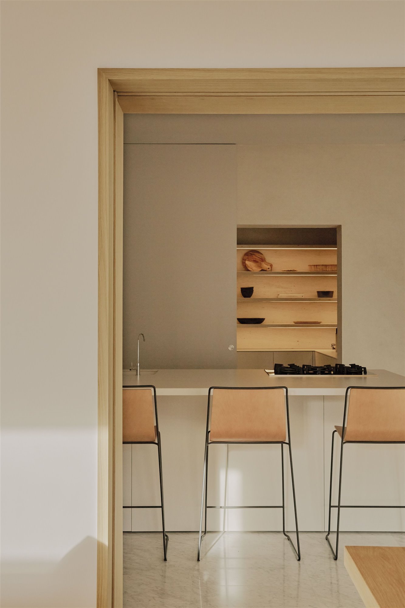 cocina minimalista beige con taburetes de madera, isla blanca y estantería con platos