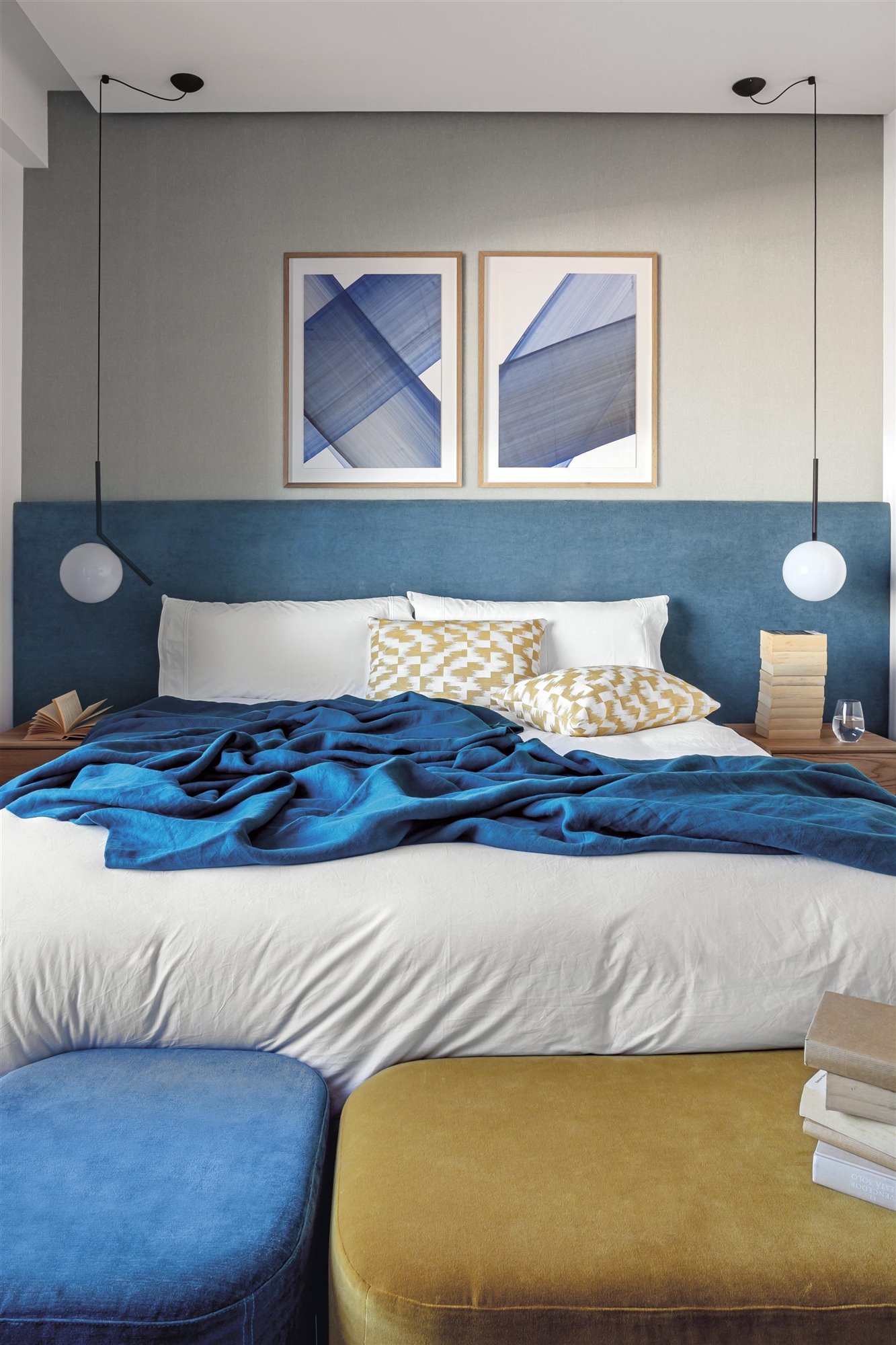 dormitorio  azul y amarillo con cuadros, cama con cojines y lámparas de lectura redondas