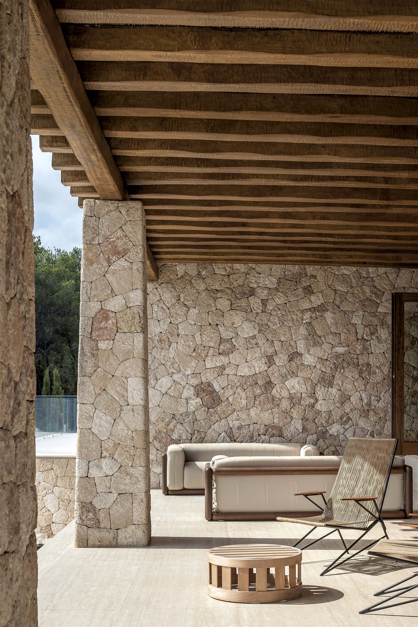 porche de casa mediterránea con techo de madera y pared de piedra, hamaca de fibra natural y sofá de exterior blanco