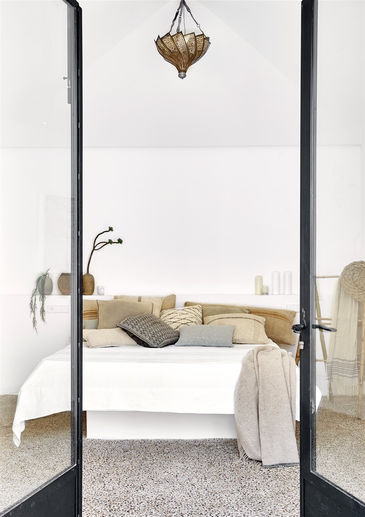dormitorio con suelo de piedras y puertas de cristal, cama con cojines y lámpara de metal