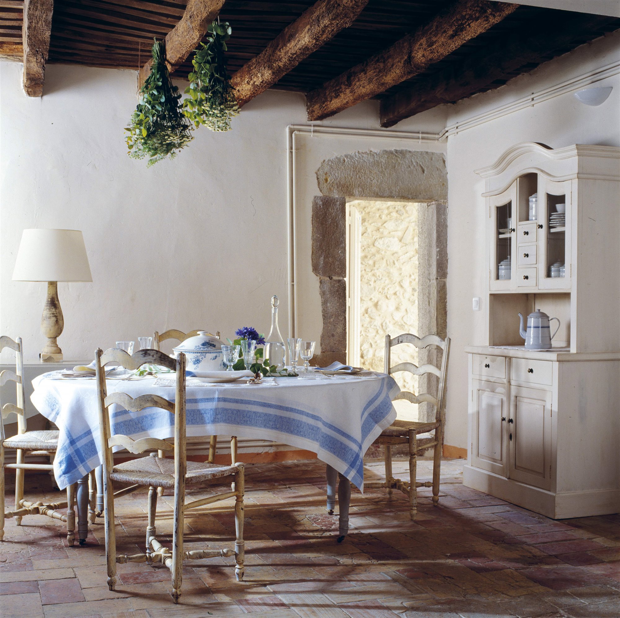 comedor rústico provenzal casa de campo con vigas de madera, mesa redonda de mantel blanco y azul, sillas envejecidas y mueble de vajilla