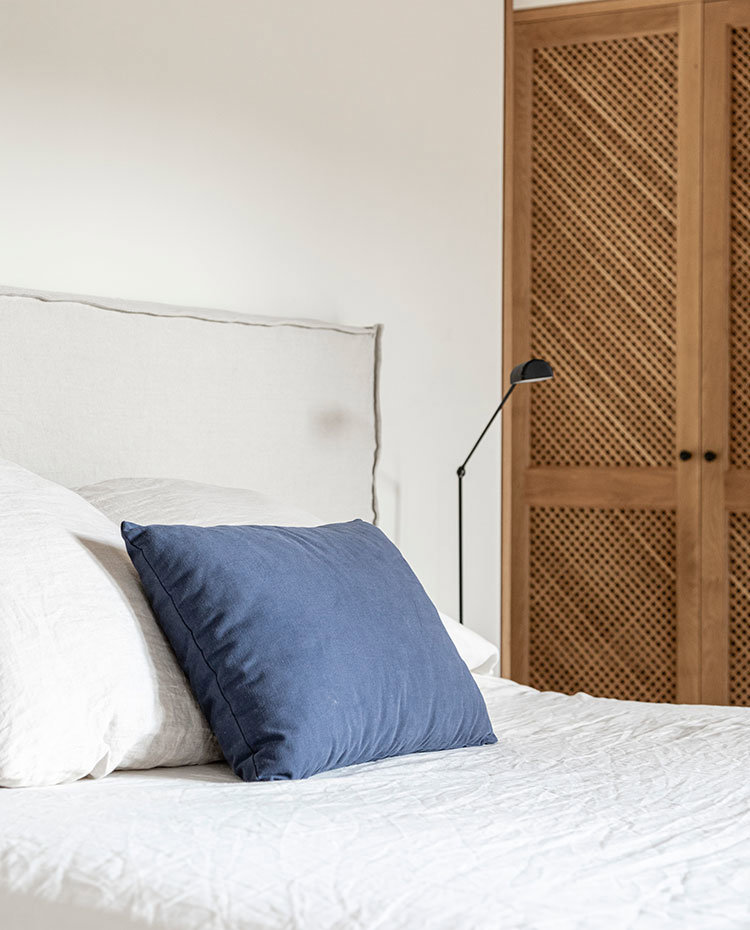 Cabecero de cama tapizado con tejido de lino, coji´n azul, luminaria de sobremesa negra y armarios con celosi´a.