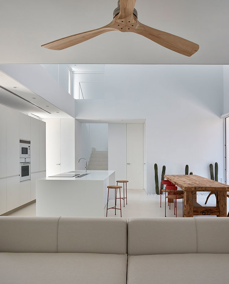 Sofa con ventilador de madera junto a cocina abierta con isla de trabajo, toda en blanco y mesa de comedor de madera