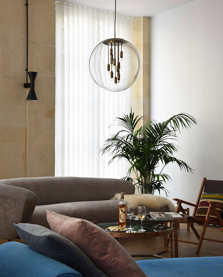 Salo´n con sofa´ marro´n, y chaise longue azu´l con cojines, dos mesitas auxilaries, silla de madera, la´mpara suspendida con patalla esfe´rica y planta junto a ventana, aplique de pared en negro