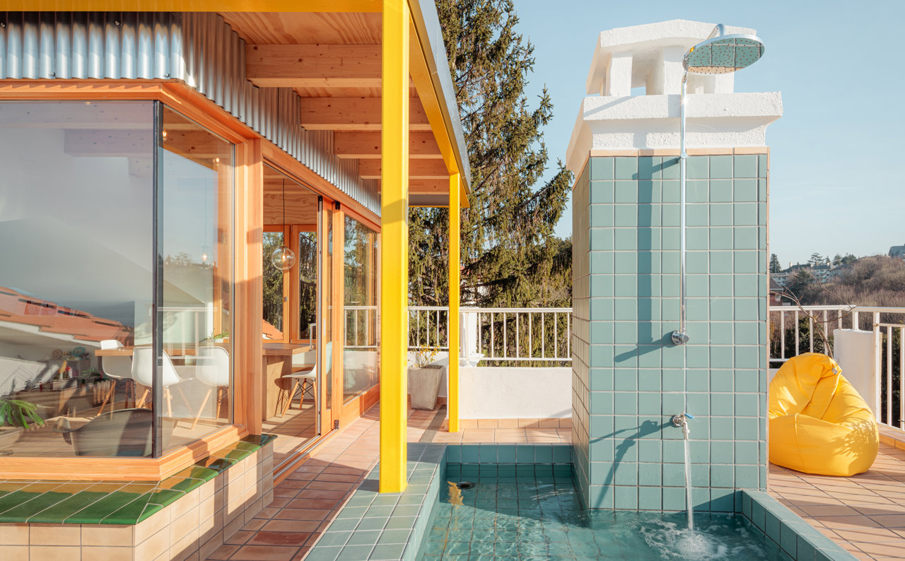 Un pabellón de madera y vidrio se abre al exterior, donde baldosas de colores y una sugerente ducha crean una atmósfera lúdica. 