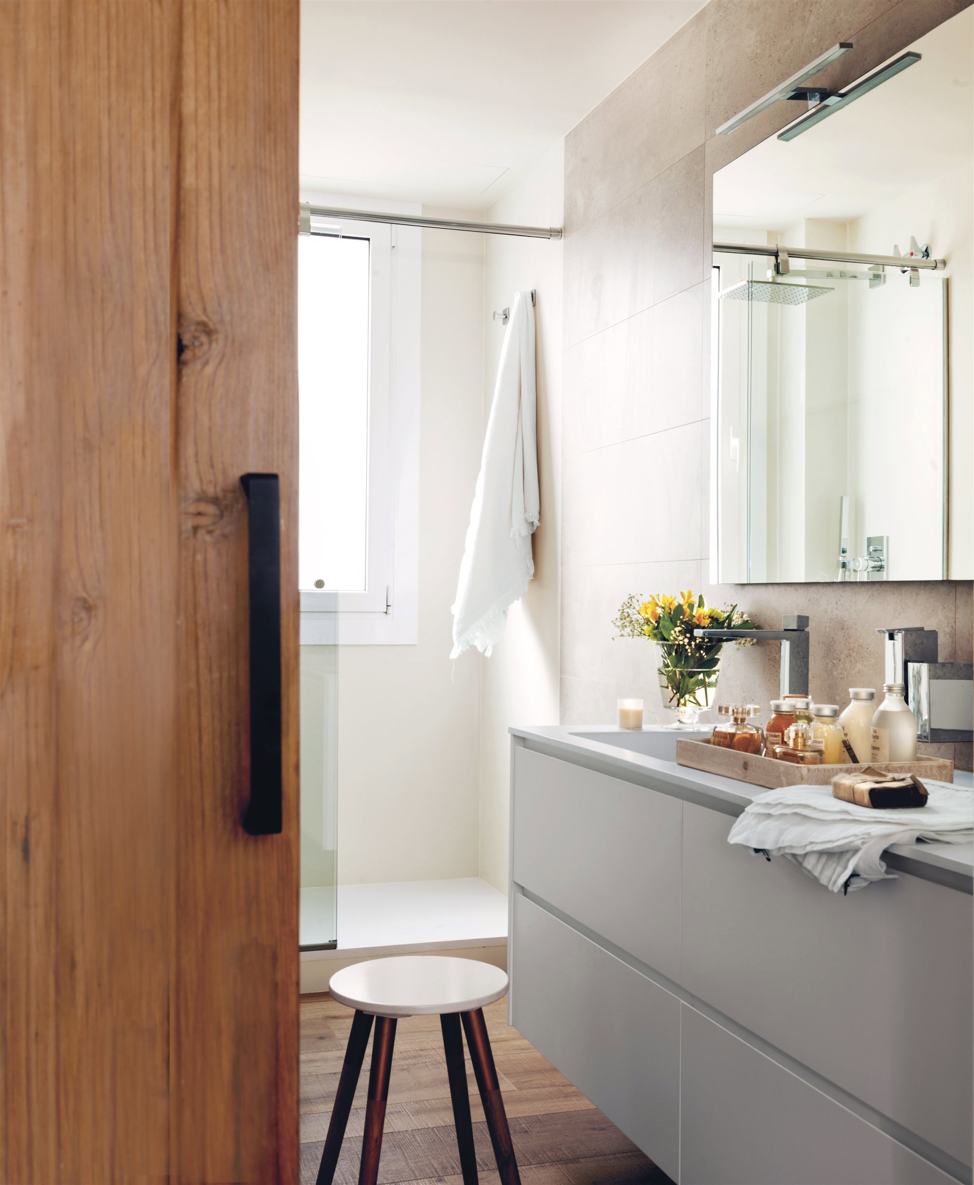 baño con puerta corredera de madera, ducha, mueble gris, taburete de madera y flores