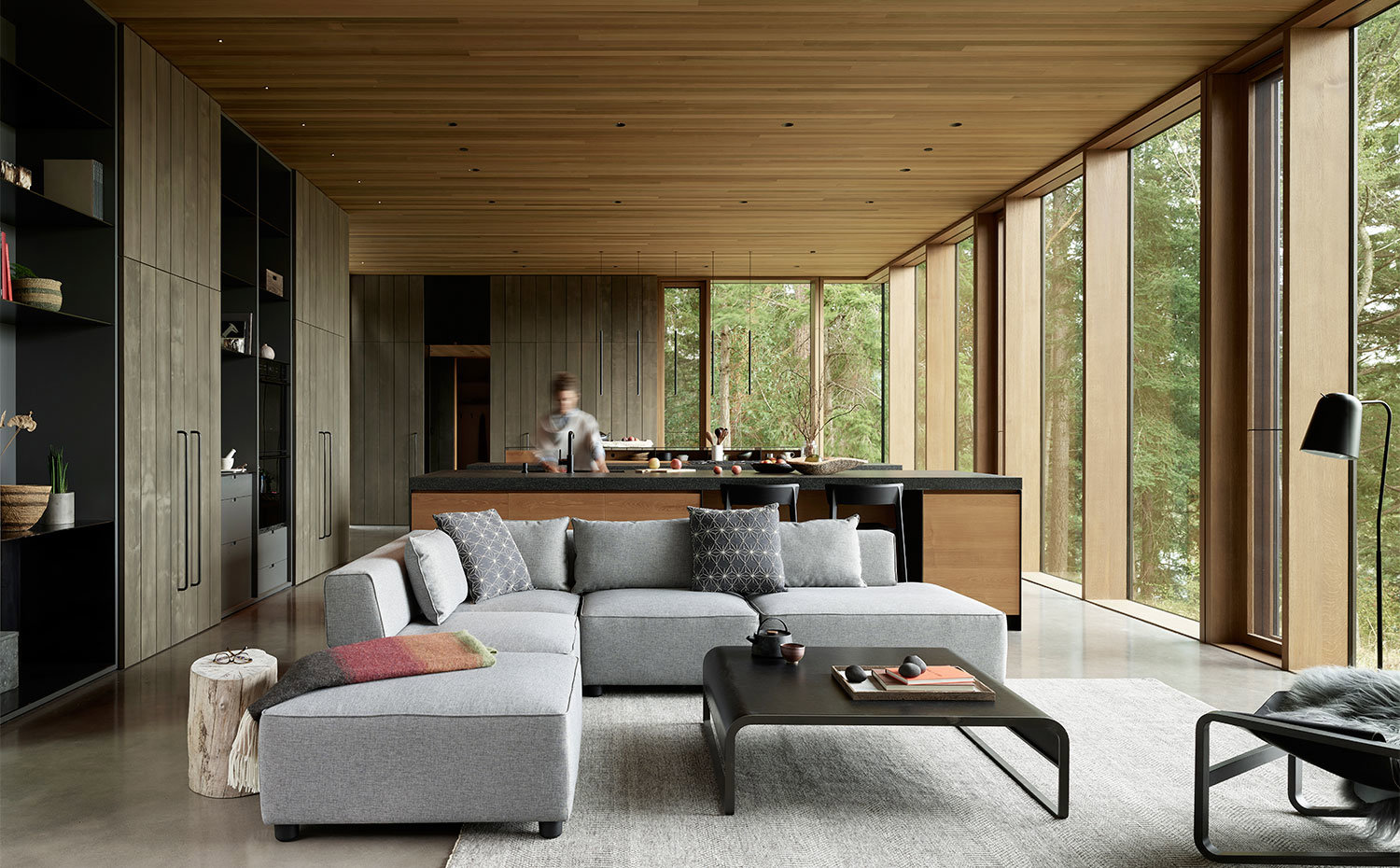 Salón moderno con cocina abierta, sofá en gris, mesa de color negro y mobiliario y techo revestido de madera.